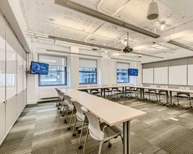 A modern conference room features long tables arranged in a U-shape, with screens mounted on opposite walls and ample overhead lighting.