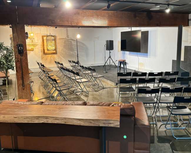 A spacious room with rows of folding chairs arranged facing a screen, accompanied by a couch made of leather and exposed wooden beams.