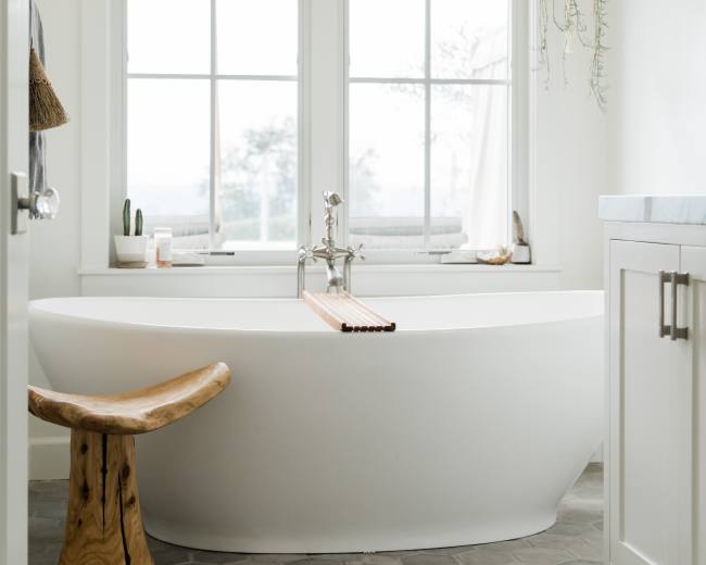 A modern bathroom features a freestanding white tub beside a large window and a wooden stool on a hexagonal stone floor.