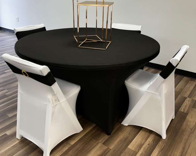 A round black table is set in a room with four white chairs adorned with black sashes, and a gold geometric centerpiece is positioned on the table.