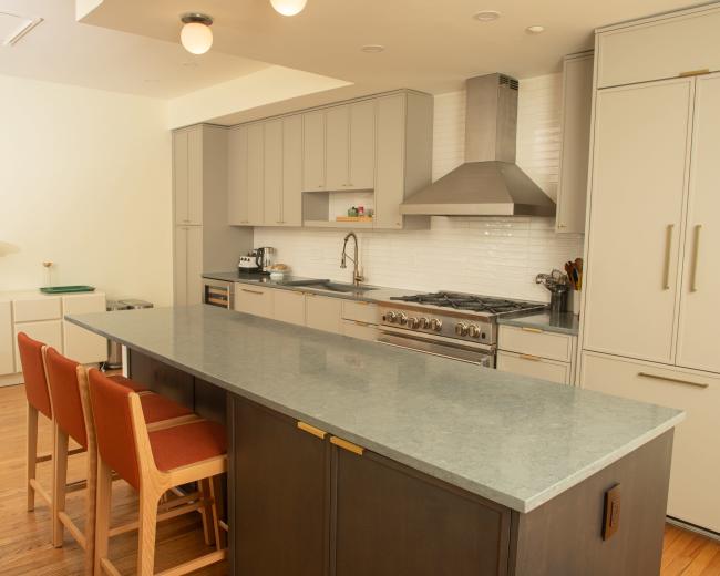The image shows a modern kitchen with gray cabinetry, a large countertop island with three wooden stools, and stainless steel appliances.