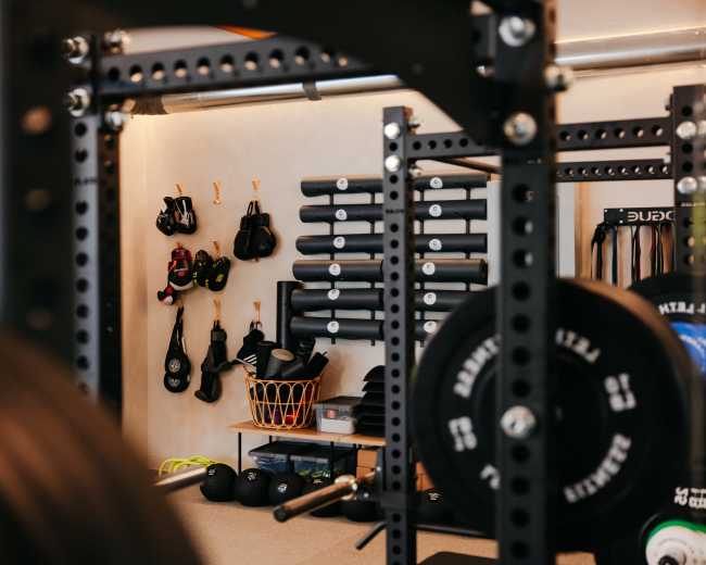 The image shows a gym setting with a weightlifting rack in the foreground and workout equipment, including boxing gloves and exercise mats, neatly organized on the wall behind.
