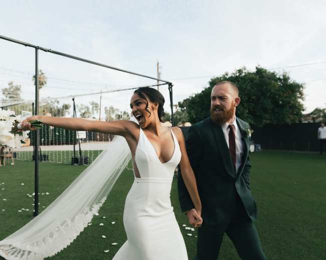 A couple celebrates their wedding by holding hands and smiling as they walk together in an outdoor venue.