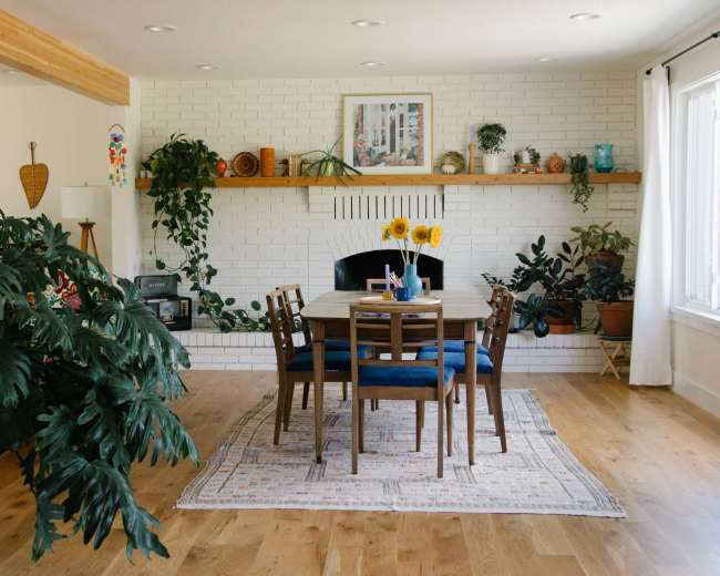 A dining area features a wooden table surrounded by chairs, with a fireplace and plants adorning the white brick wall.