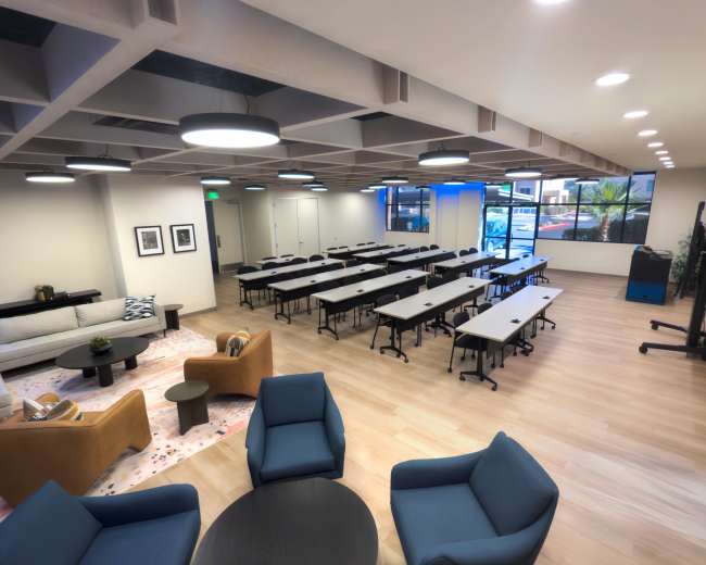 The image shows a modern classroom or meeting space with rows of tables and chairs arranged for a gathering, along with a lounge area featuring sofas and a coffee table.