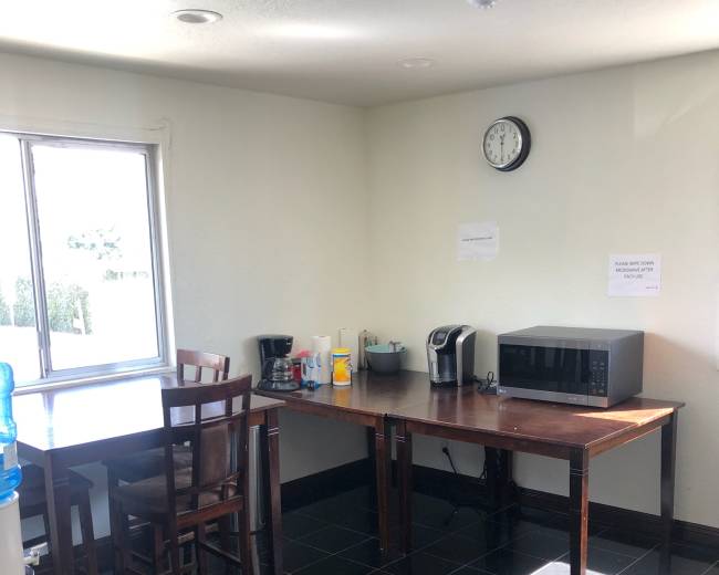 A small kitchen area features a table with chairs, a coffee maker, a microwave, and a water cooler along with a window and wall clock.