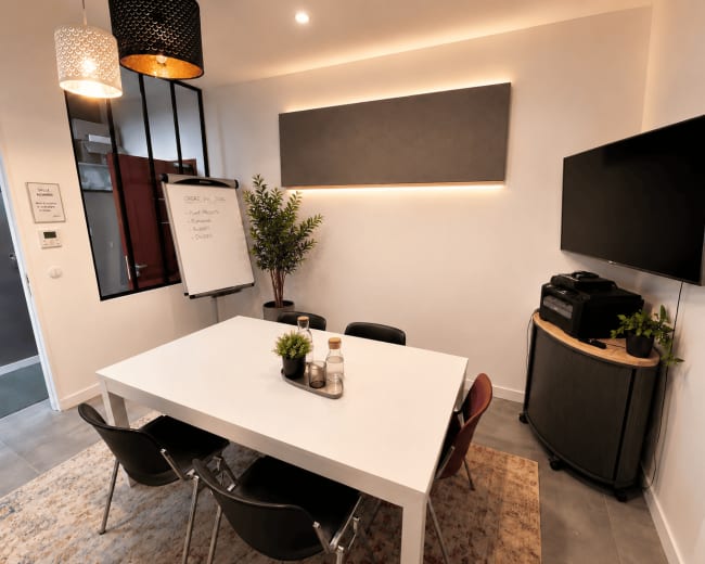A modern meeting room features a white table surrounded by black chairs, a wall-mounted TV, a plant, and a whiteboard illuminated by pendant lights.