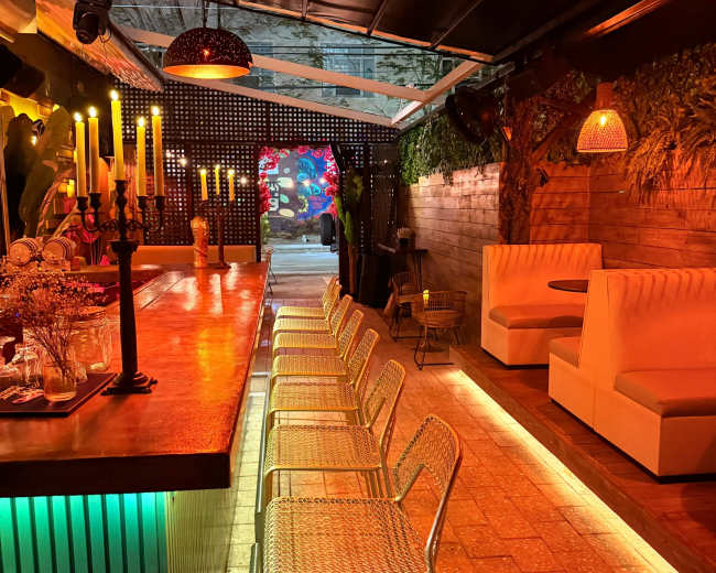 The image shows a dimly lit bar area with a long counter, several metal chairs, and cozy seating booths in a modern design, accented by colorful ambient lighting.