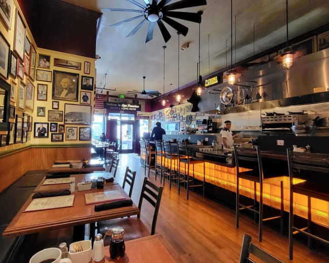 The image shows the interior of a restaurant with wooden tables, a bar area, and walls adorned with framed photographs.