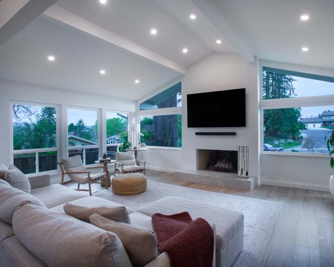 The image shows a modern living room with a light-colored sofa, two armchairs, a coffee table, and a large television mounted on the wall, surrounded by large windows offering views of the outside.