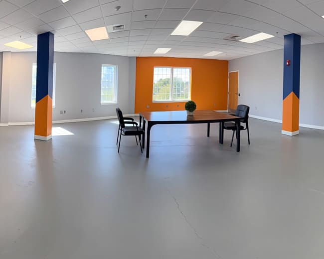The image shows a spacious, empty office interior with gray floors, orange accent walls, and a table surrounded by chairs.