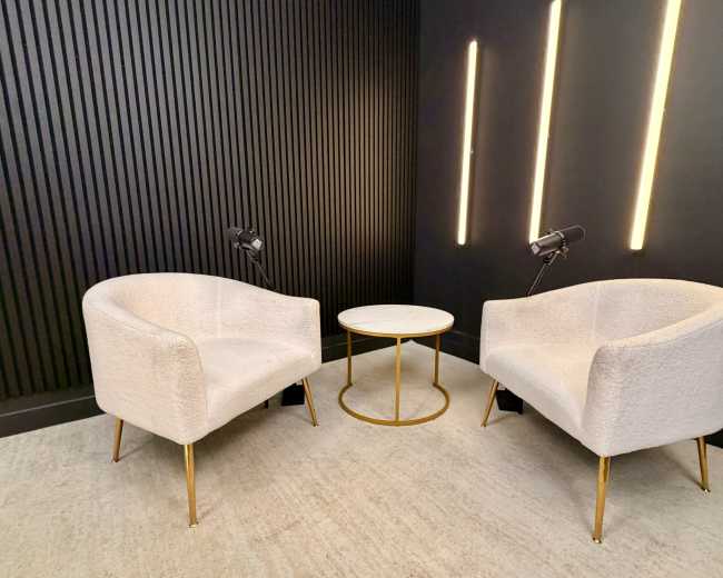 Two upholstered chairs with gold legs are positioned across from each other next to a round table, against a dark wall featuring vertical paneling and modern lighting.