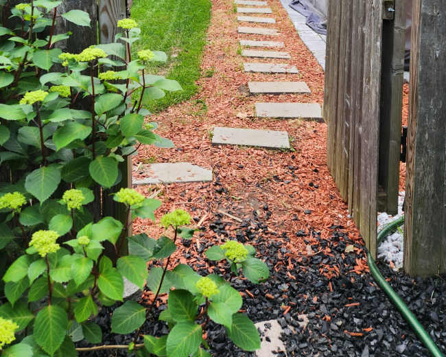 A stone pathway lined with mulch leads through a wooden gate into a well-maintained yard with greenery and outdoor furniture.