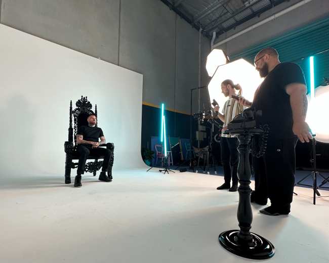A man sits on an ornate throne in a studio setting while crew members adjust lighting and equipment around him.