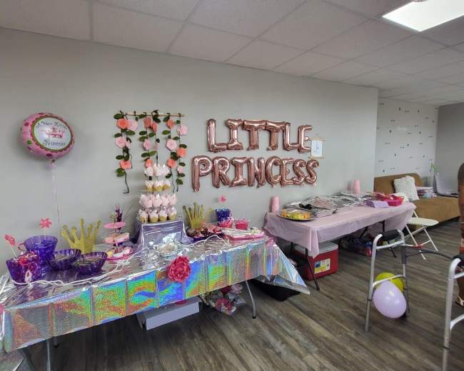 A party setup with a table featuring a pink tablecloth, a tiered cupcake stand, and decorations that read "LITTLE PRINCESS" along the wall.