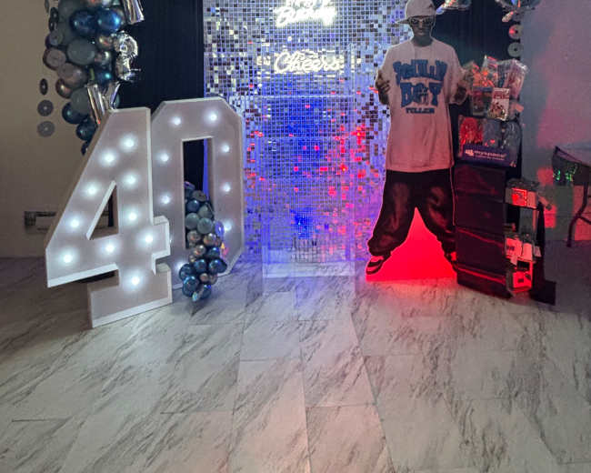 The image shows a decorative setup for a 40th birthday celebration, featuring a large "40" made of lights, a backdrop of mirrored tiles, balloons in silver and blue, and a figure dressed in a T-shirt with a party theme.
