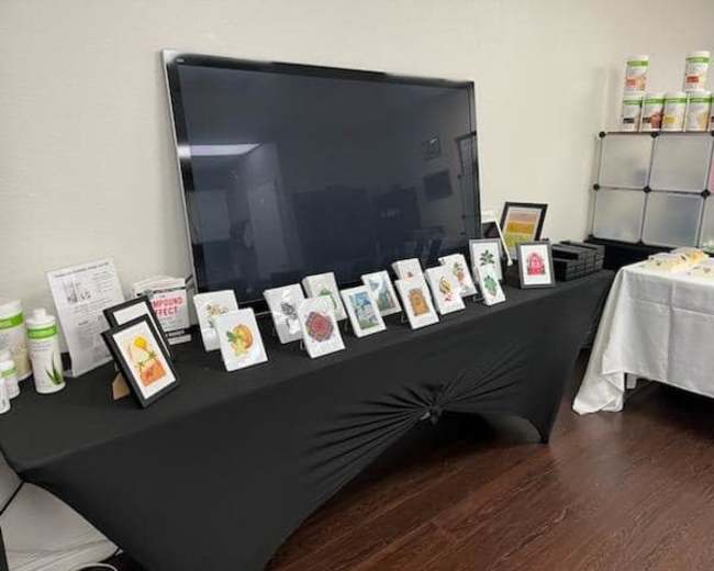 A table with a black tablecloth displays framed artwork and a television screen in a well-lit room.