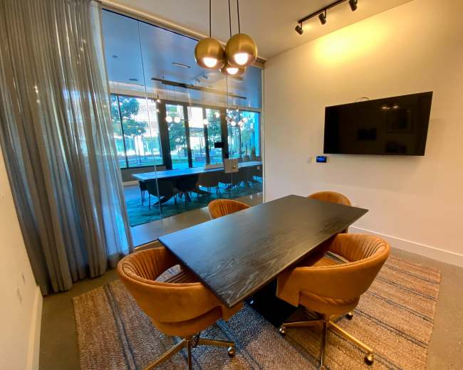 A conference room features a rectangular table surrounded by four chairs, with a television mounted on the wall and large glass doors leading to an outdoor area.