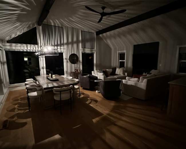 A dimly lit living space with a large dining table, various seating arrangements, and shadows created by overhead lighting on the walls and ceiling.