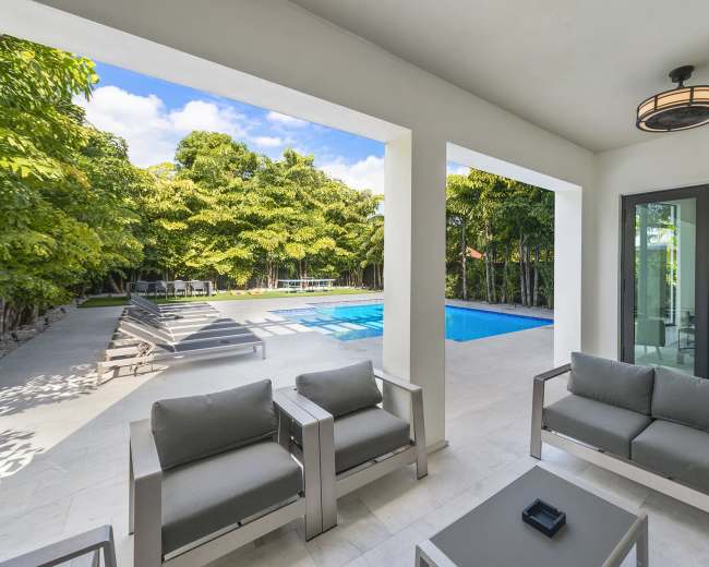 The scene shows a modern outdoor lounge area featuring gray seating and a glass table, with a swimming pool and sun loungers in the background surrounded by greenery.