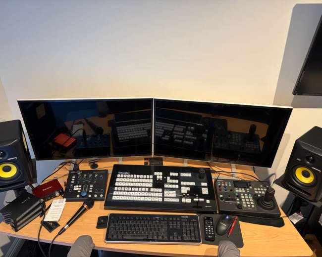 The image shows a digital editing workstation featuring two large monitors, a control panel with buttons and dials, a keyboard, speakers, and various tools organized on a wooden desk.