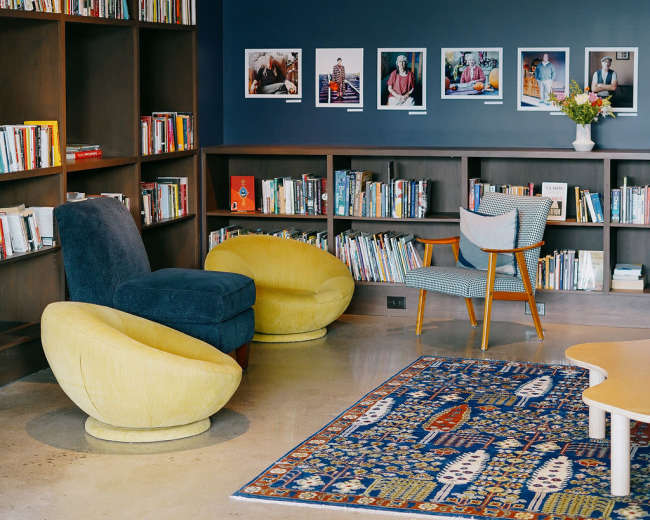 The scene features a cozy reading area with two armchairs, a bookshelf filled with books, framed photographs on the wall, and a colorful area rug.