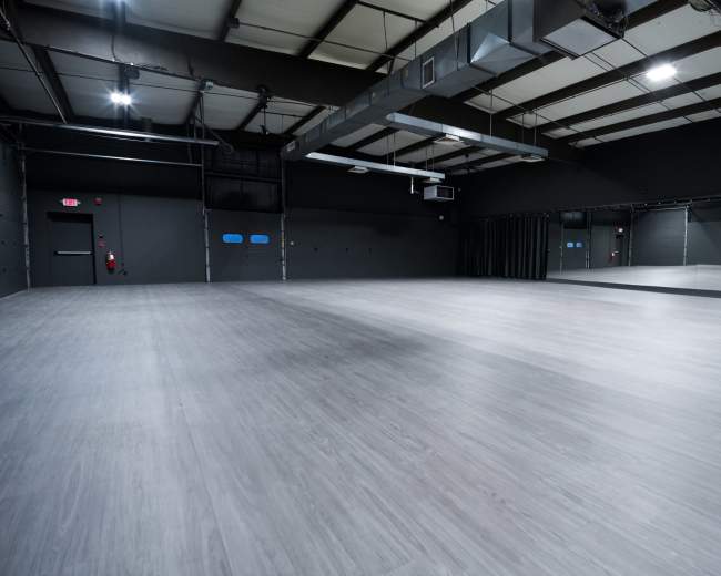 The image shows a spacious, empty dance studio with gray flooring and mirrors along one wall.