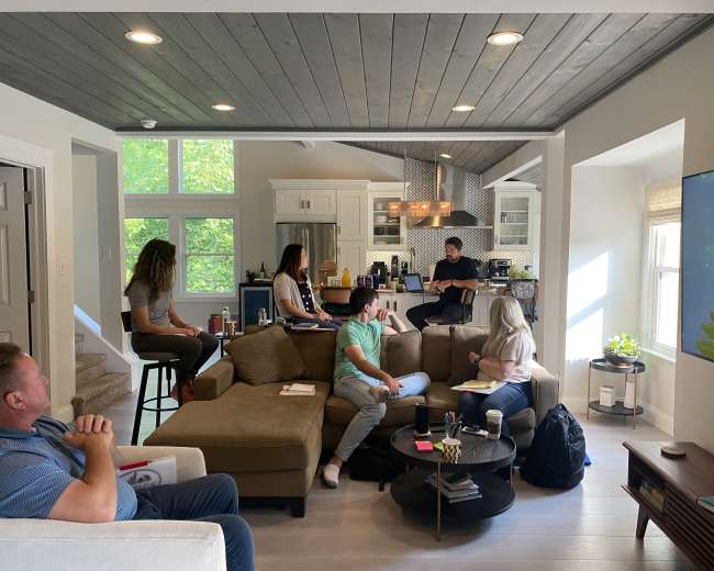 A group of six people is seated in a living room, engaged in a discussion while a kitchen area is visible in the background.
