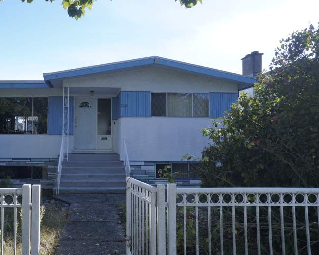 The image shows a beige-colored, single-story house with blue accents and a front porch, surrounded by overgrown shrubs and a white fence.