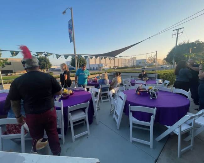 A group of people gathers around purple tablecloth-covered tables in an outdoor setting during an event.