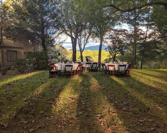 A row of tables with white tablecloths is set up on a grassy area under trees, overlooking a mountainous landscape.