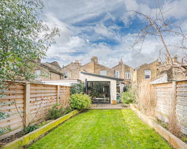The image shows a landscaped garden with neatly trimmed grass, bordered by wooden fences and featuring a contemporary glass extension of a house in the background.
