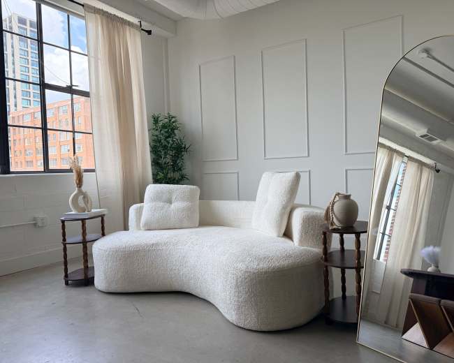 The image shows a modern, minimalist living space featuring a white, curvilinear couch, two small side tables, a large mirror, and a plant near a window with sheer curtains.