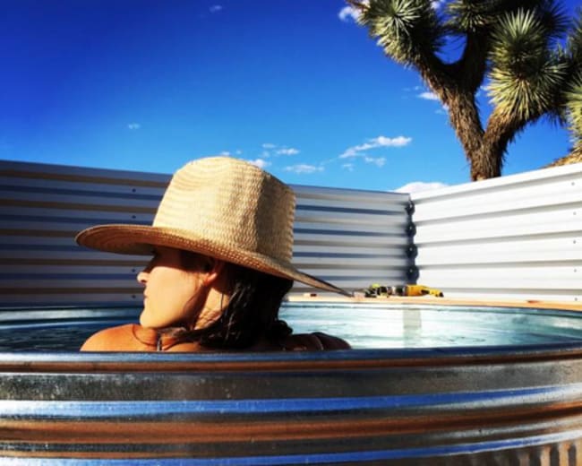 A person wearing a straw hat relaxes in a metal hot tub against a backdrop of clear blue sky and a Joshua tree.