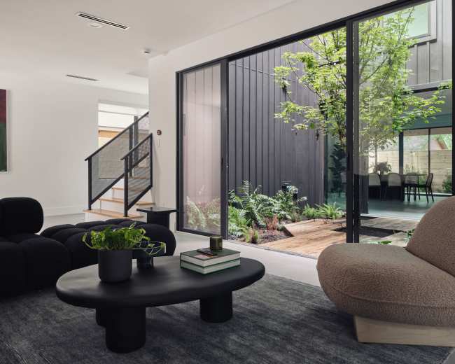 A modern living room features a black coffee table, plush seating, and large glass doors that open to a landscaped outdoor area.