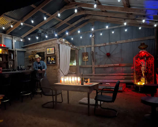 The interior of a rustic cabin is illuminated by string lights, featuring a bar area and a table with candles, while a person stands near the bar.