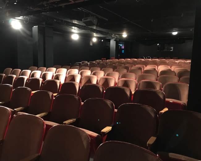 The image shows a dimly lit theater with rows of red-upholstered seats, arranged in a symmetrical pattern.