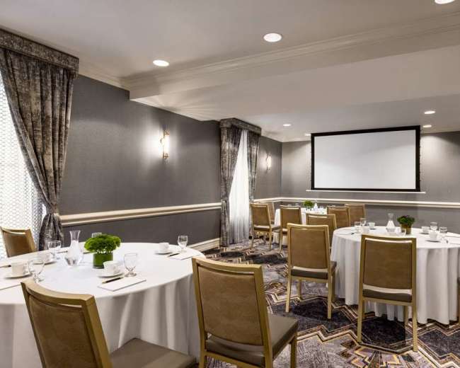 A well-lit meeting room features round tables set for dining, with a large projector screen at the front and elegant curtains at the windows.