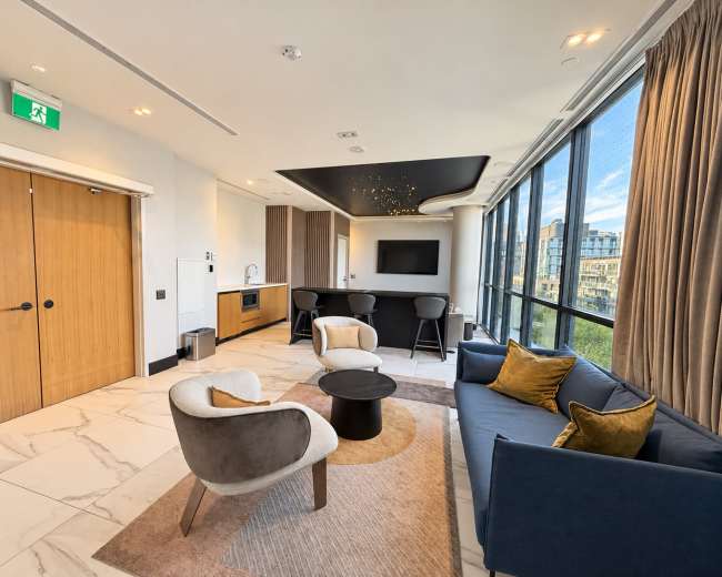 The image shows a modern living room with two armchairs, a sofa, a round coffee table, and large windows overlooking a cityscape.