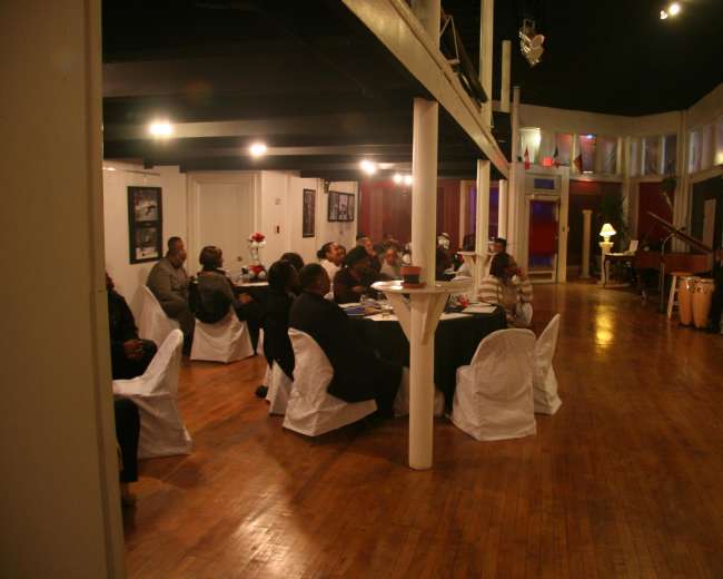 A group of people sits at tables in a dimly lit art space listening to a performer on stage.