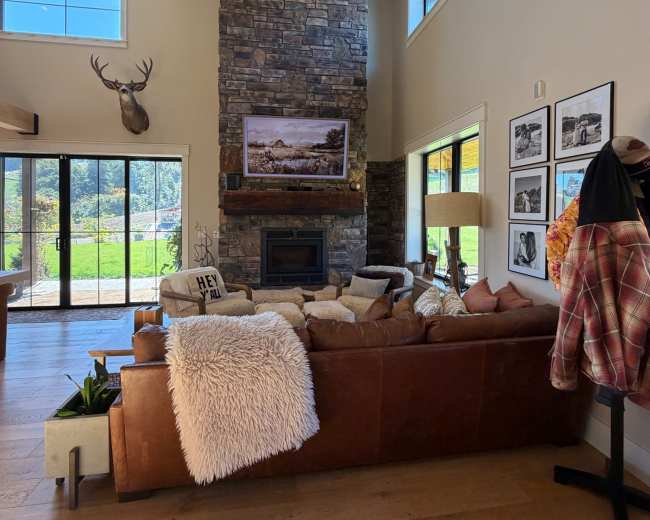 The image shows a modern living room featuring a large stone fireplace, a leather sofa adorned with pillows, and a coffee table with a small plant, all set against a backdrop of large windows that offer a view of greenery outside.