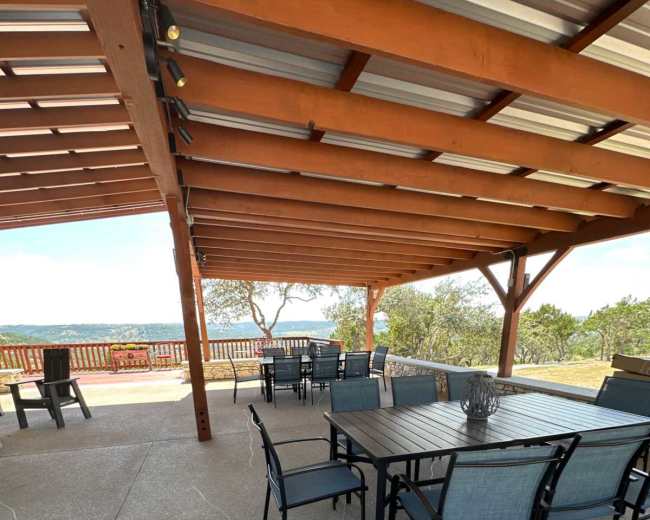 The image shows a covered patio area with several tables and chairs arranged on a concrete surface, overlooking a landscape with trees and a distant view.