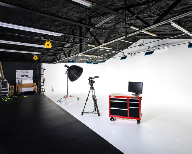 The image shows a photography studio with a camera on a tripod, a large light umbrella, a red tool cart, and a monitor against a white backdrop.