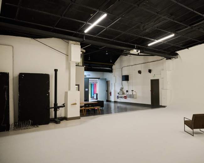 The image shows a spacious, minimalist studio with a single chair positioned on a white floor, surrounded by contrasting dark walls and empty spaces.