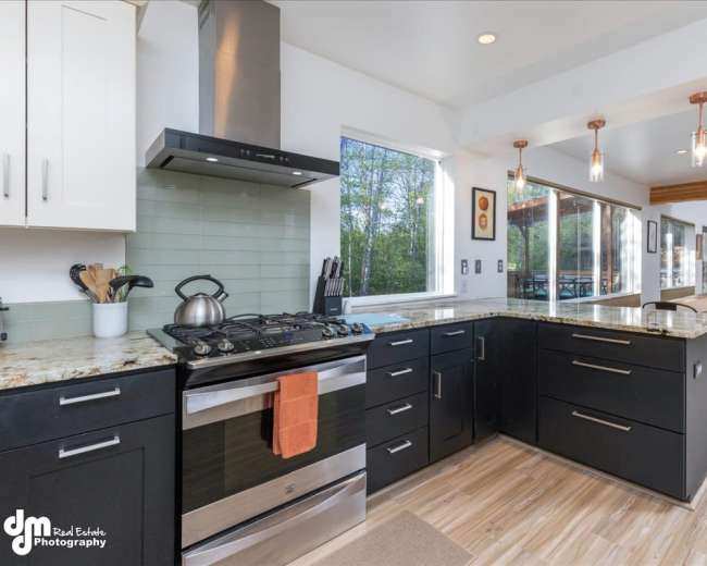 A modern kitchen with black cabinets, a stainless steel stove, and a granite countertop, adjacent to a dining area with pendant lighting.