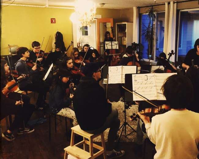 A group of musicians is seated in a studio, playing string instruments while reading sheet music.