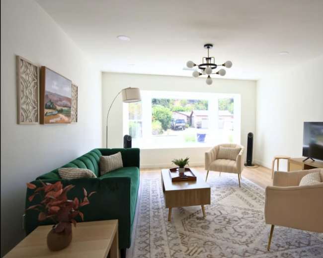 The image shows a modern living room with a green sofa, beige armchairs, a wooden coffee table, and a television against a white wall, featuring a large window that illuminates the space.