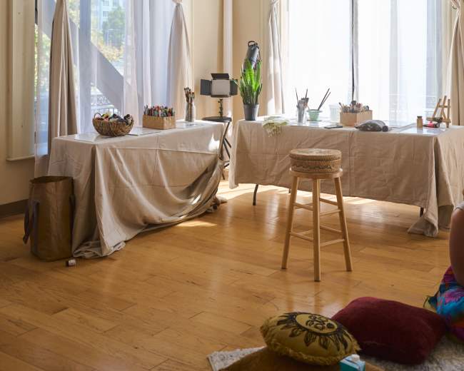 The scene depicts an artist's studio with two tables covered in white fabric, natural light streaming through large windows adorned with white curtains, and various art supplies arranged on the tables.