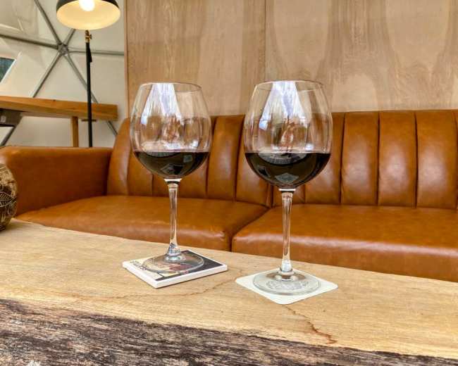 Two glasses of red wine are placed on coasters atop a wooden table in front of a brown leather couch.