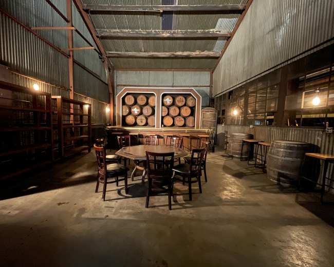A dimly lit room features a wooden table surrounded by chairs, with shelves and barrels against the corrugated metal walls.